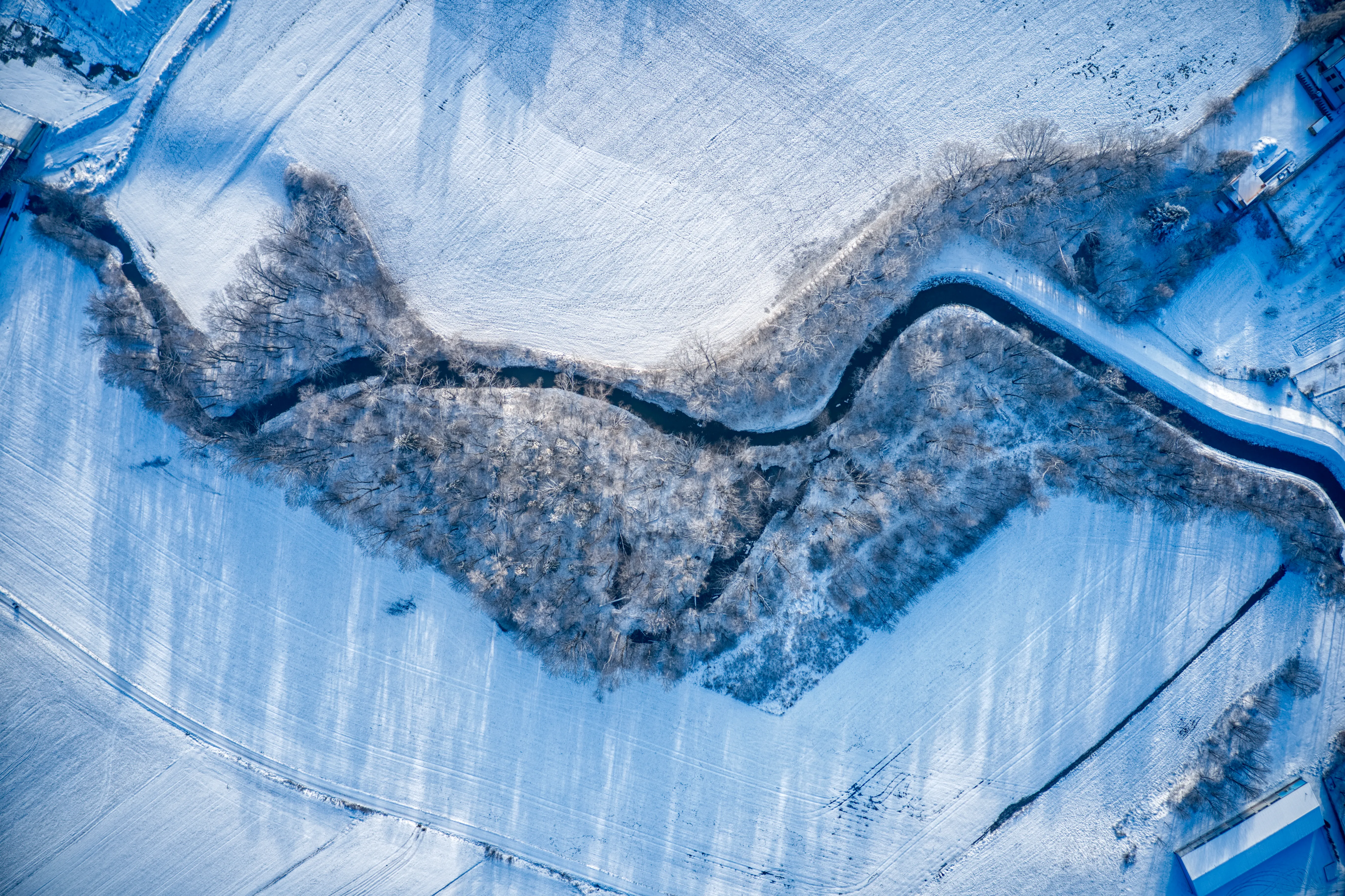 Image captured by a drone camera of wintery snowy fields with a river meandering through the middle